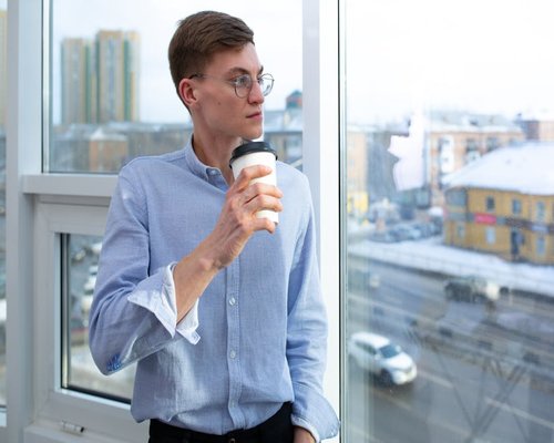 Person looking out of a high rise office window holding a warm cup of coffee, looking away from screen